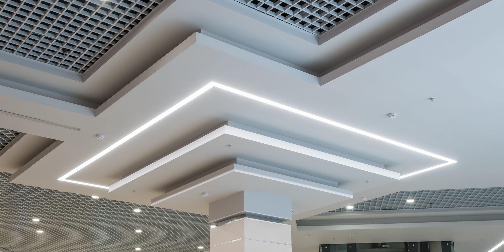 A stepped white ceiling with a continuous LED strip running around its angled edges, between round downlights and metal grid panels