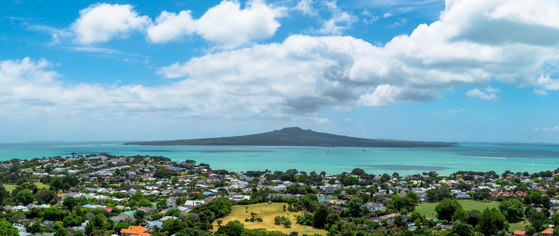 Suburban rooftops and trees running down to turquoise harbour water, with Rangitoto Island on the horizon under a bright, cloudy sky
