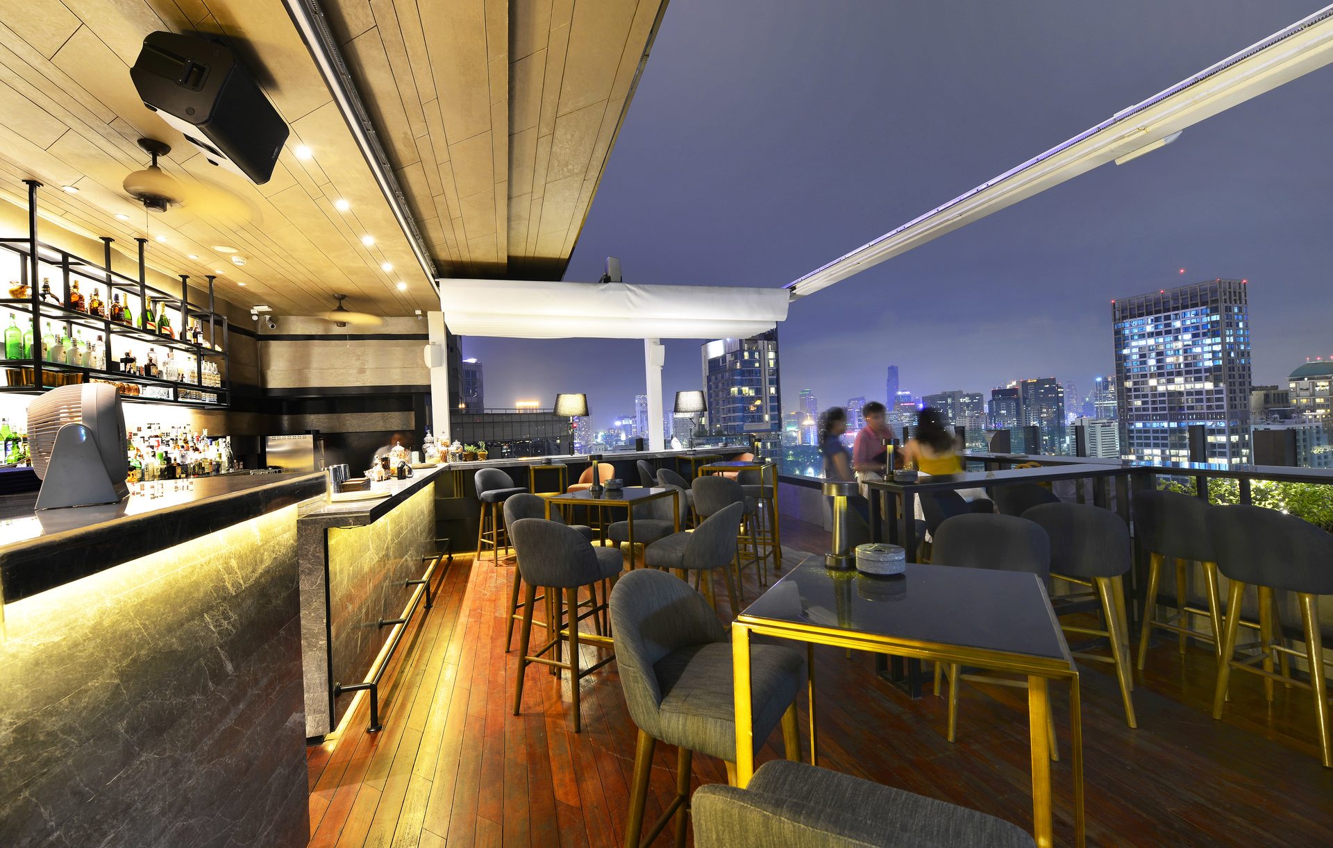 A rooftop bar at night with high stools, warm lighting under the counter and lit city towers beyond the railing