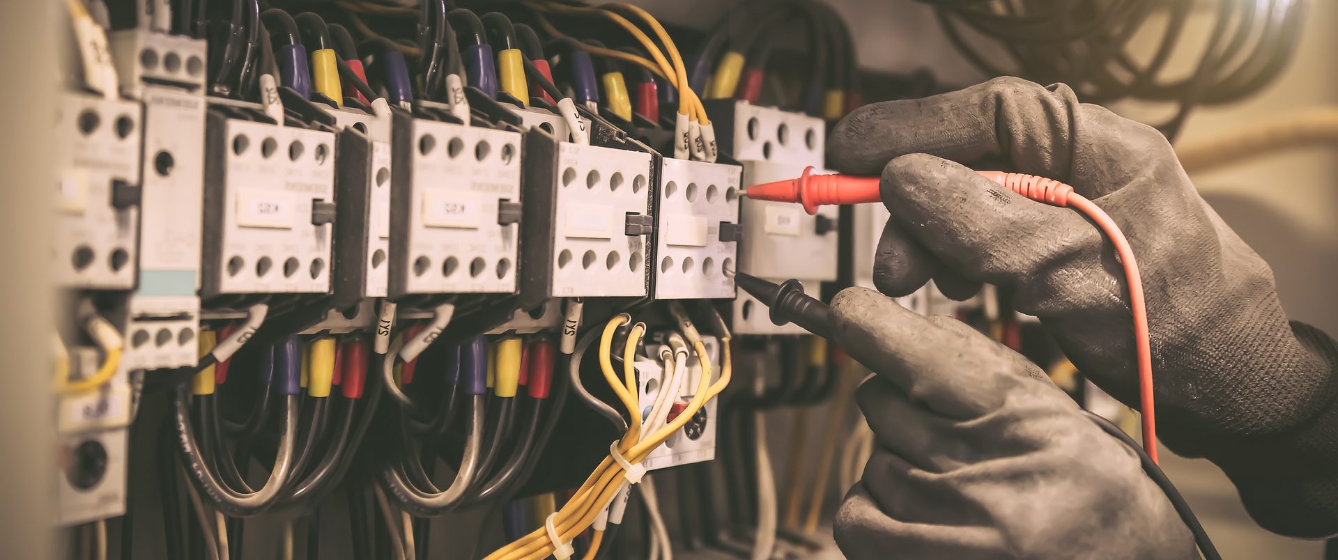 A gloved hand holding a red test probe against a row of wired contactors inside an electrical panel